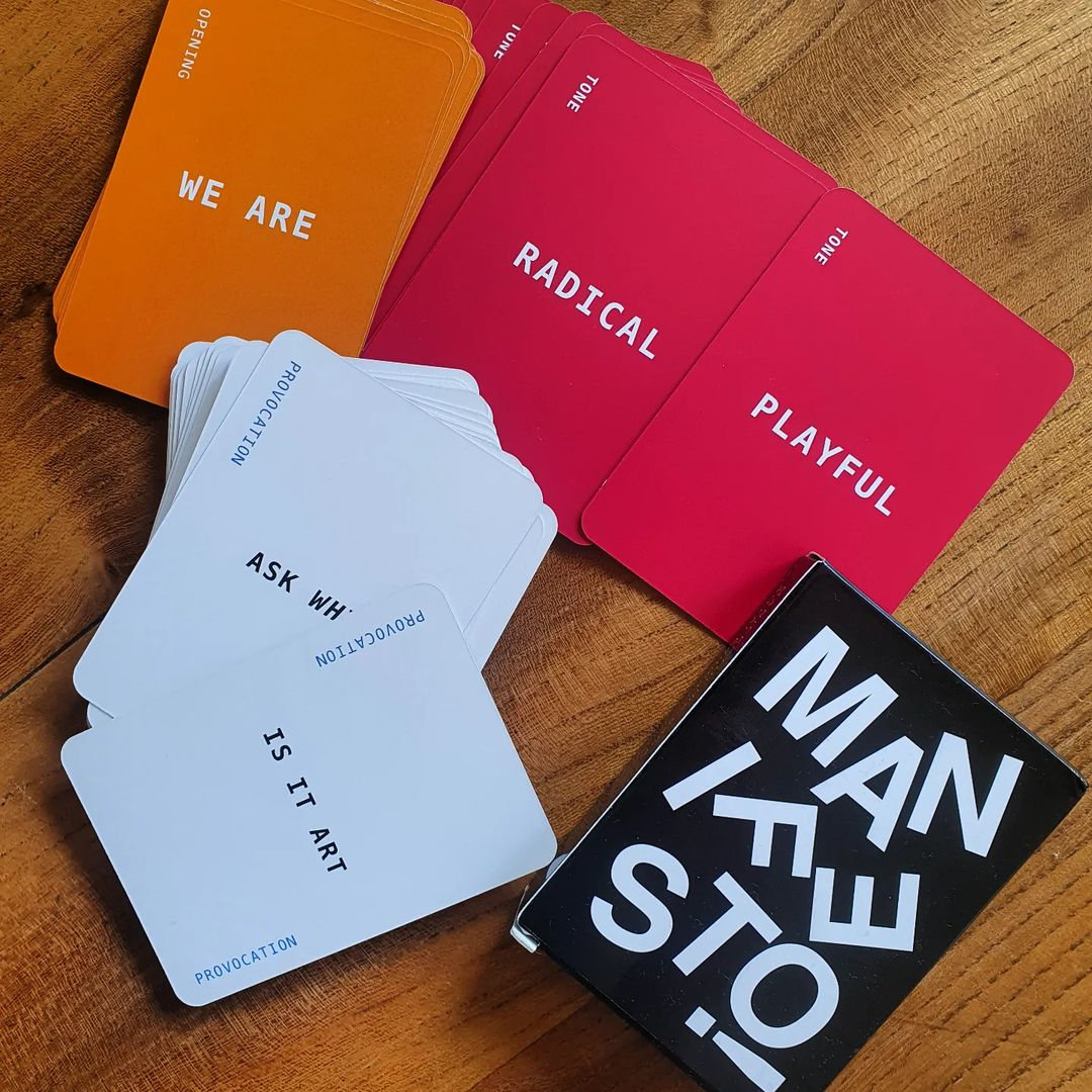 Photo of the MANIFESTO! card game on a desk. In addition to a bold, black box with white lettering, we see cards of assorted colors (red, orange, and white) reflecting different suits: Tone, Opening, and Provocation.