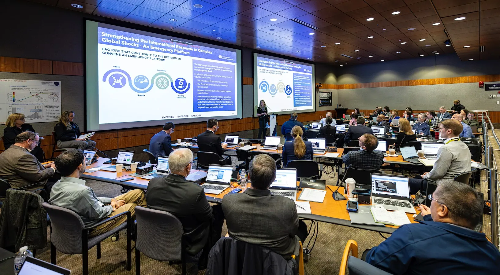 Photo of many people in a large room, all seated, and looking at screens in a slide presentation.