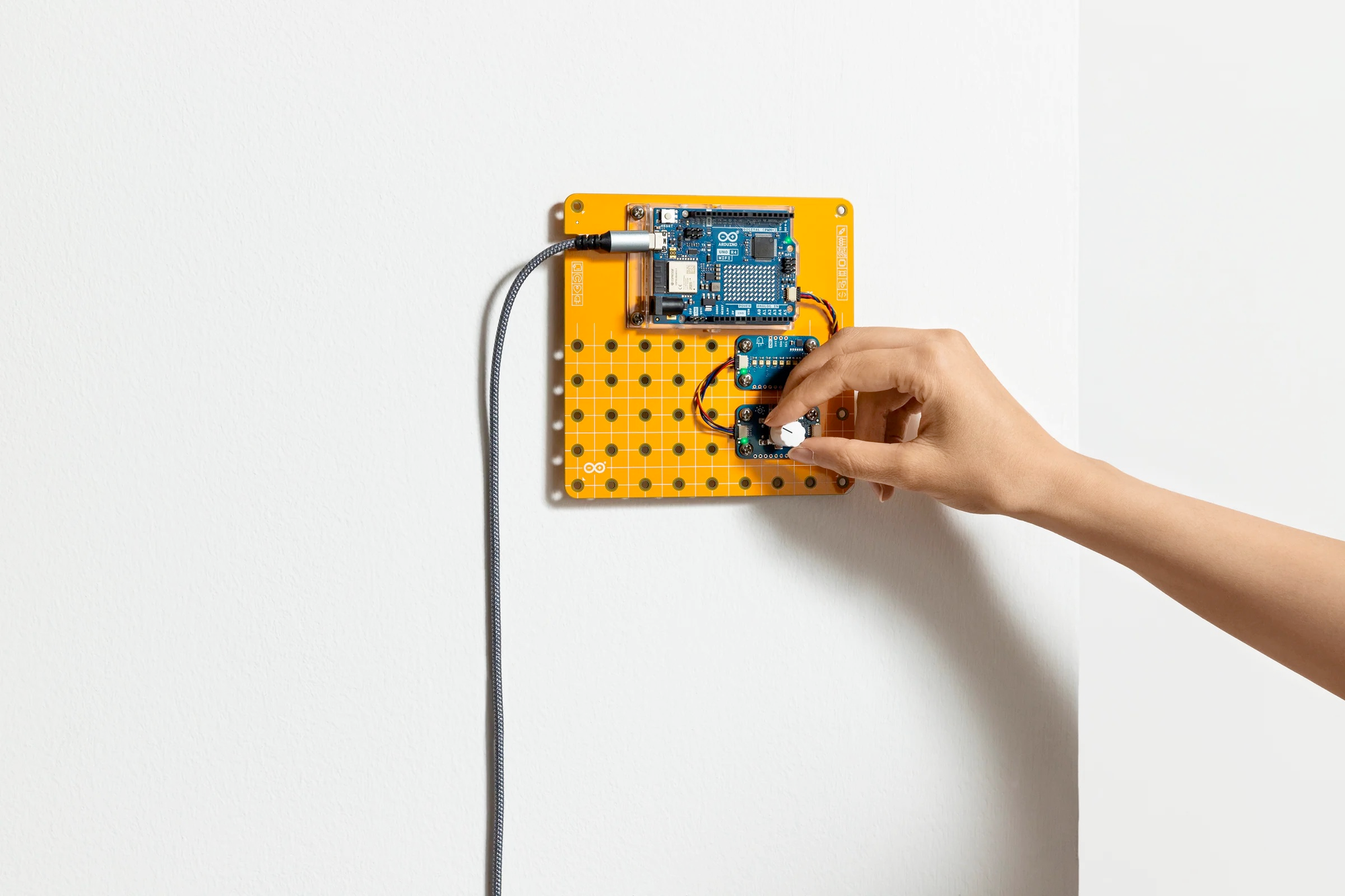 A hand adjusting a button or knob on an Arduino plug and make kit, mounted to a white wall on a yellow bread-board-like backing.