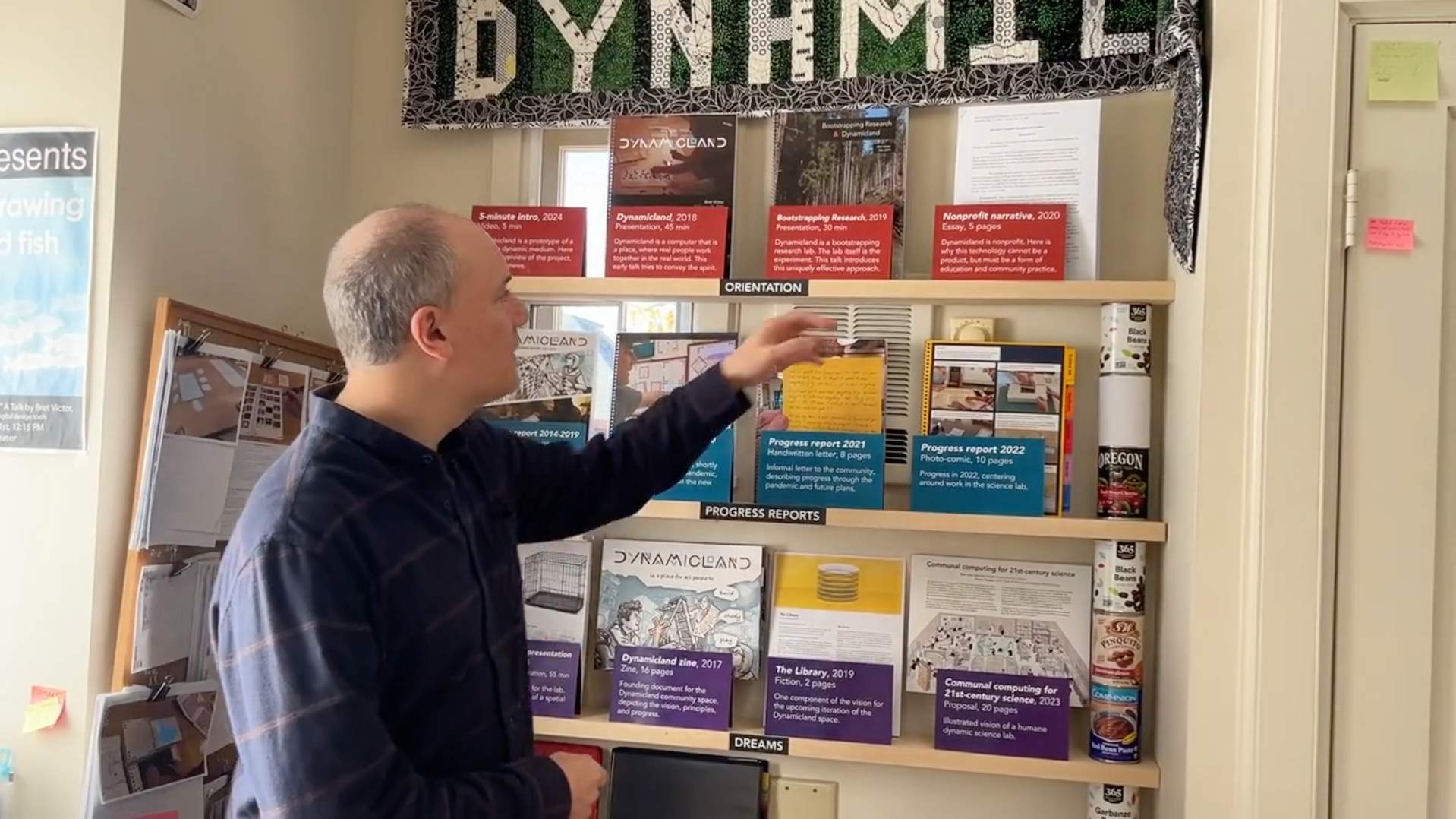 Photo of a person standing in front of stacked shelves, with spiral-bound books facing out, and placards of different colors in front of each book.