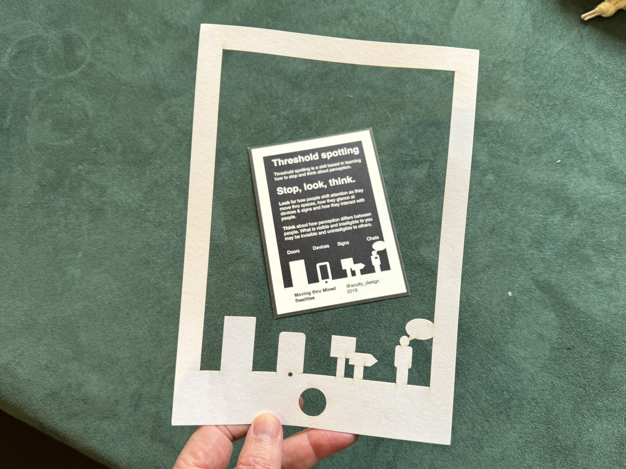 Photo of a hand holding an A5 sized sheet of paper, with a window cutout that turns the page into a frame or window onto other things. Silhouetted in this frame are what looks like a door, a mobile device, signs, and a person talking. Beyond the view on a table are instructions for using this frame.