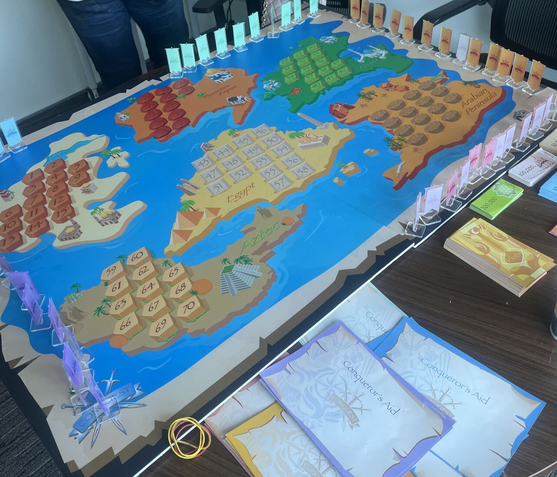 Photo of a game board, depicting a fictional map. There are six islands, each covered in a series of roughly a dozen numbers. Assorted standees, cards, player aids, money, and similar game materials surround the board.