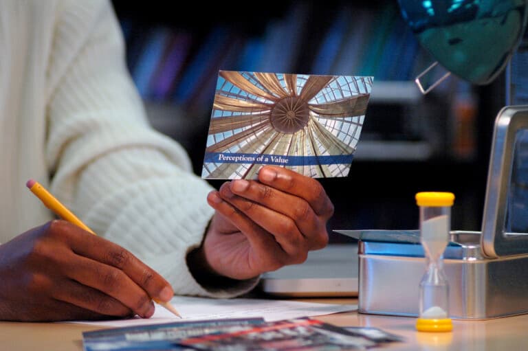 Close up photo of a person at a desk. One hand holds a pencil while the other holds a single card, upon which we see an abstract photo and the text "Perceptions of a Value."