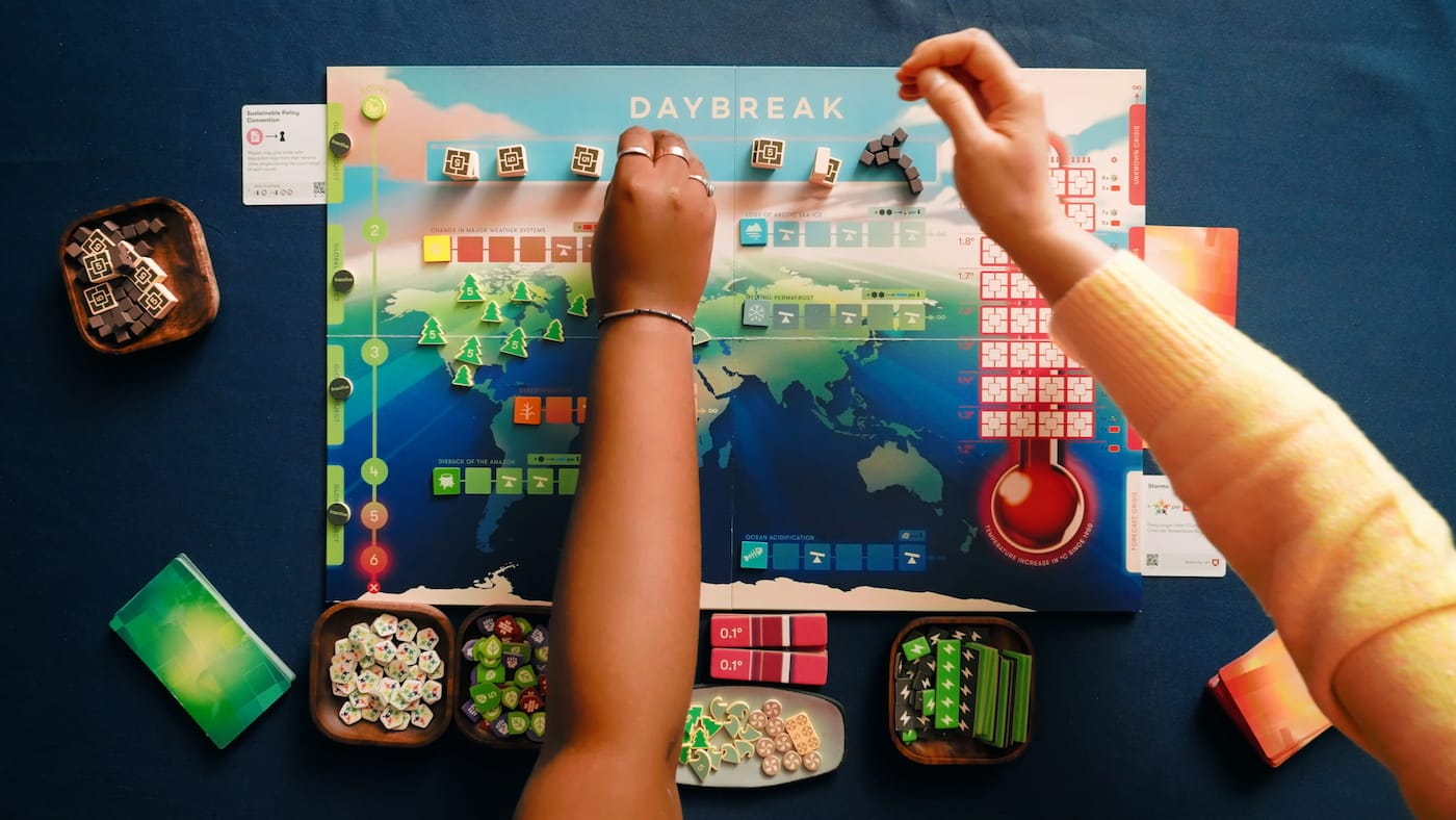 Bird-eye view photo of people (we can only see their arms) playing the board game Daybreak.