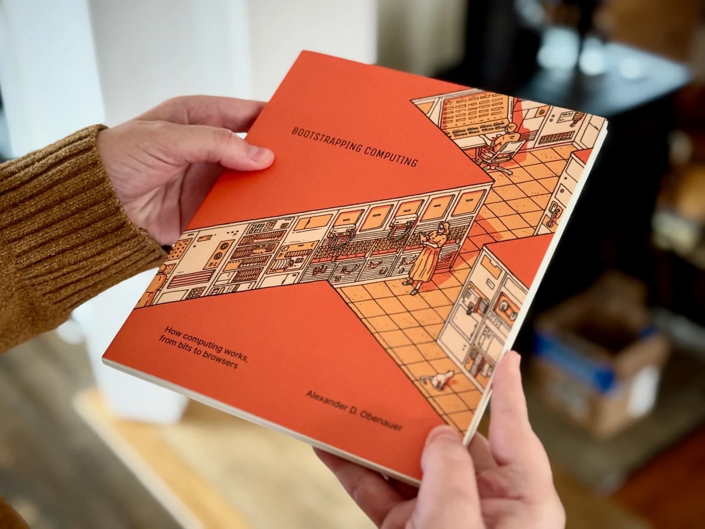 Close up photo of a pair of hands holding a book: "Bootstrapping Computing: How computing works from bits to browsers" by Alexander D. Obenauer.