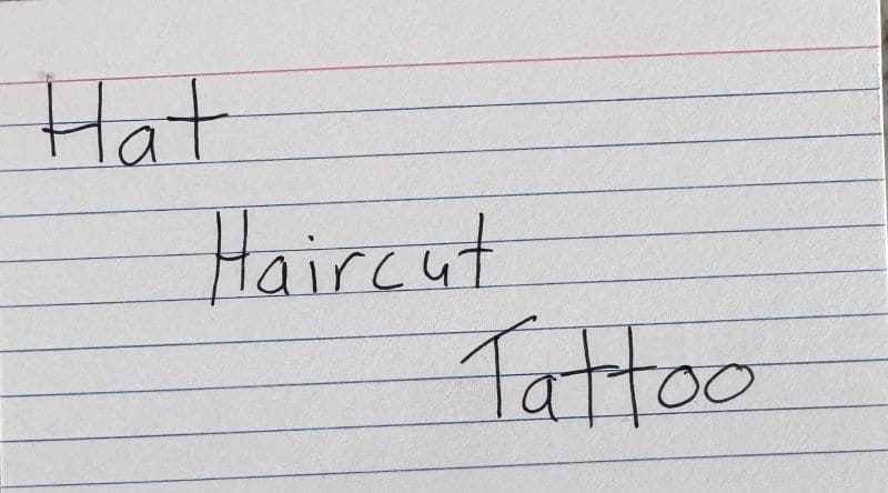 Photo of an index card with the words "Hat," "Haircut," and "Tattoo" written on it.