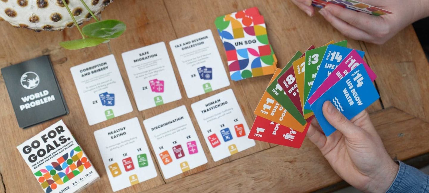 An overhead view of the 'Go for Goals' card game in progress on a wooden table. A hand holds a fan of colorful UN Sustainable Development Goals (SDG) cards showing Goals 1 through 14, including 'NO POVERTY' (red), 'GOOD HEALTH' (green), 'LIFE BELOW WATER' (blue), and others in their distinctive SDG colors. On the table are laid out several white game cards including 'WORLD PROBLEM' (dark gray), 'CORRUPTION AND BRIBERY,' 'SAFE MIGRATION,' 'TAX AND EXTREME COLLECTION,' 'HEALTHY EATING,' 'DISCRIMINATION,' and 'HUMAN TRAFFICKING,' each with colored SDG icons and point values. A 'UN SDG' reference card with the colorful SDG wheel design sits among the cards. A small succulent plant and another player's hand holding cards are visible at the edges of the frame.