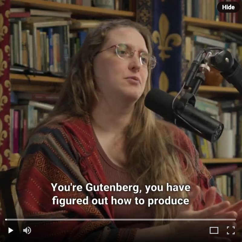 A video screenshot showing a person seated in front of bookshelves filled with books and a blue banner with gold fleur-de-lis patterns. A microphone on a boom arm is visible to the right. White subtitle text at the bottom reads 'You're Gutenberg, you have figured out how to produce…' Video playback controls are visible at the bottom of the frame.