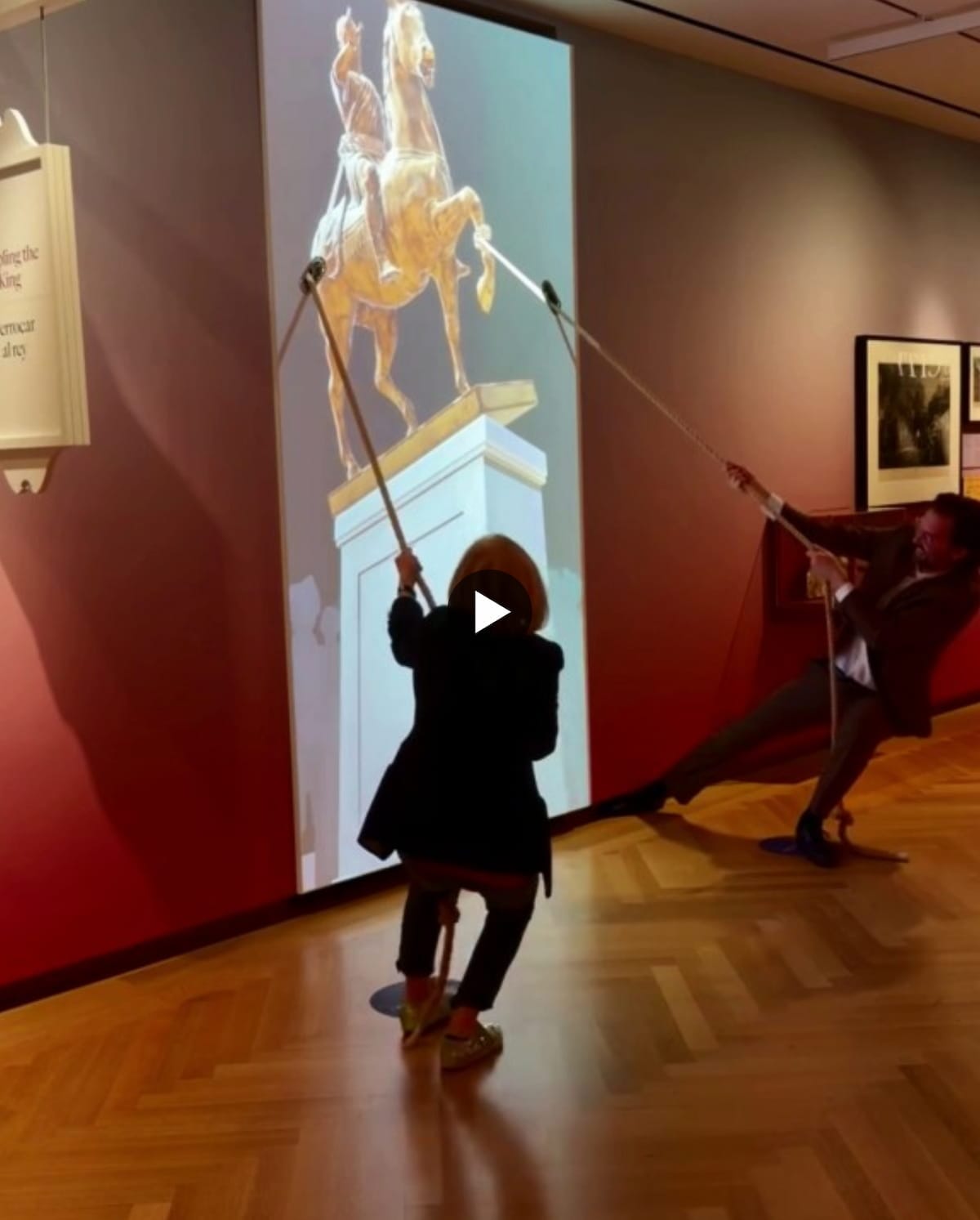 A video screenshot showing an interactive museum installation where two people pull on ropes to topple a projected statue of King George III. The projection displays an animated golden statue of the king on horseback atop a white pedestal against a light blue background. The scene is set in a museum gallery with hardwood floors, red walls, and framed artwork visible. A white play button overlay indicates this is a paused video. One person in dark clothing stands in silhouette in the foreground pulling a rope, while another person is visible in the background on the right side of the frame also pulling. The installation appears to recreate the historical act of pulling down the statue of King George III.