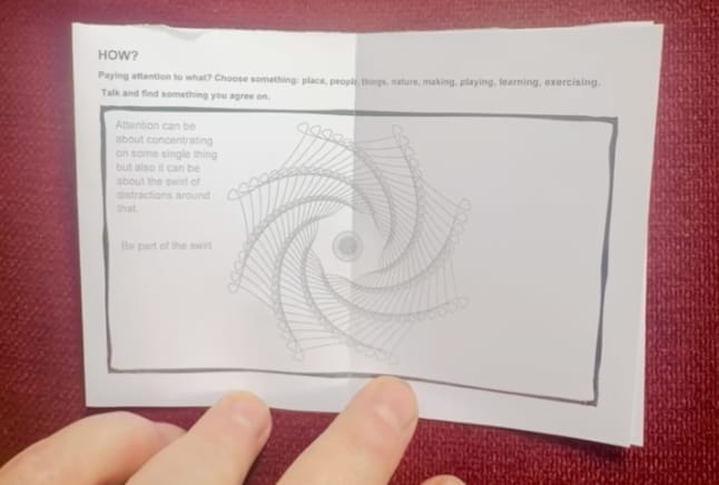 A photograph of a hand holding open a page from a zine about attention, resting on a burgundy fabric surface. The page is titled 'HOW?' and contains text explaining: 'Paying attention to what? Choose something: place, people, things, nature, making, playing, learning, exercising. Talk and find something you agree on.' Below this is a geometric spiral illustration made of intersecting straight lines creating a hypnotic rotating pattern. On the left side of the illustration is partially visible text that reads 'Attention can be about concentrating on one thing. It's focusing. But also it can be about the internal distractions around you... the part of the self.' The page appears to be from a DIY-style educational zine exploring the concept of attention.