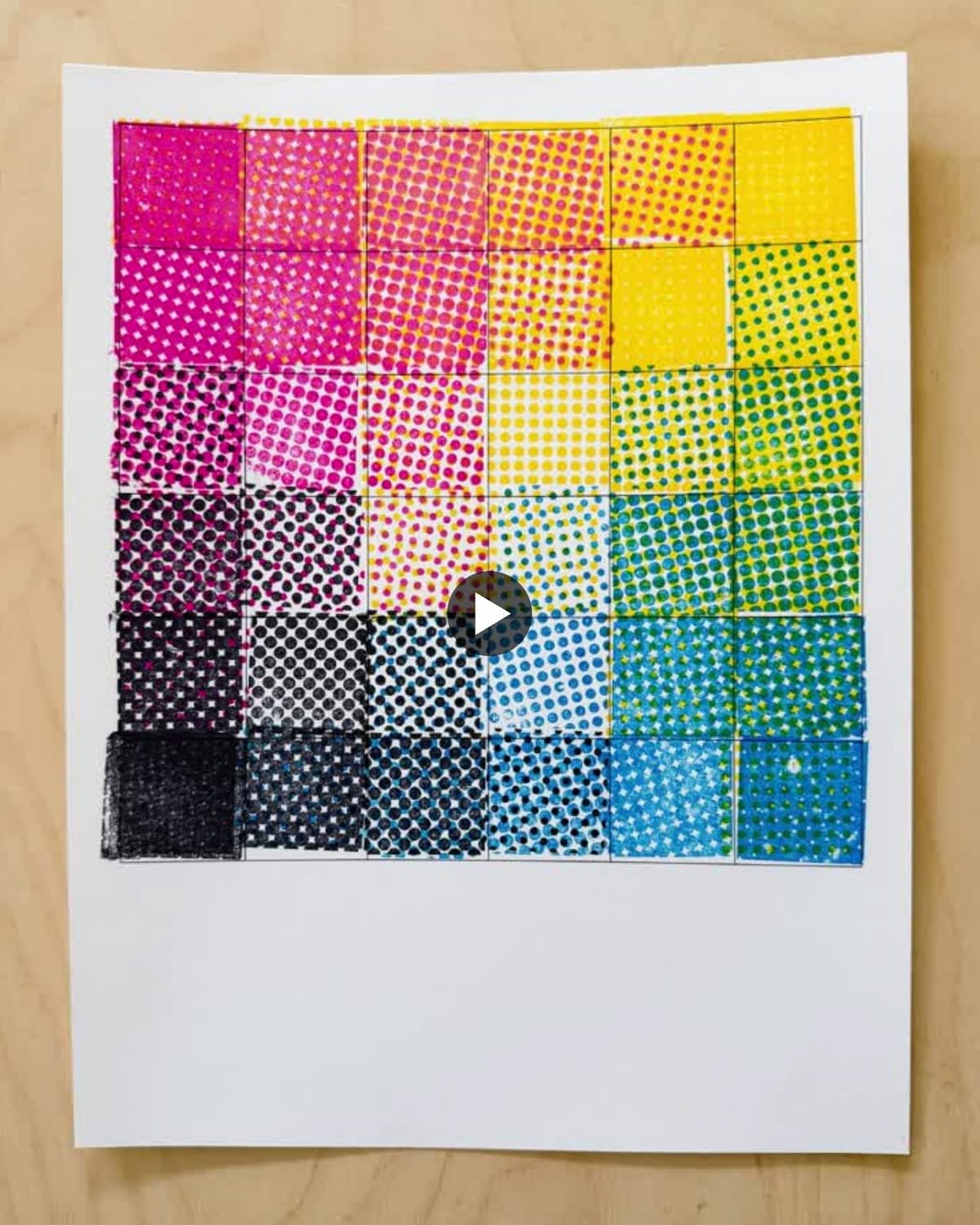 A video screenshot showing a hands-on CMYK color mixing demonstration created with stamps on white paper placed on a wooden surface. The sheet displays a 6x6 grid of hand-stamped squares showing halftone dot patterns in various colors and densities, transitioning from magenta and pink (left side) through yellow (top center) to cyan, green, and blue (right side), with black squares in the bottom left corner. Each square demonstrates different densities and overlapping patterns of stamped dots, illustrating how CMYK color mixing works in printing. A white play button overlay in the center indicates this is a paused video. This interactive exhibit piece allows people to learn about halftone screening and color theory through hands-on stamping.