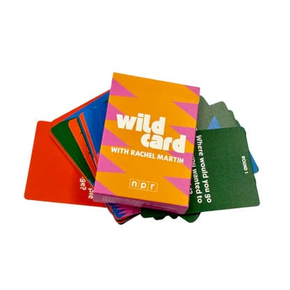 A fanned-out deck of colorful cards from the NPR podcast 'Wild Card with Rachel Martin.' The top card features an orange and pink geometric design with white text displaying the show title and NPR logo. Behind it, multiple cards in various colors including red, green, blue, and orange are visible, with one green card showing partial text reading 'Where would you go to' and 'round yourself.' The cards are conversation prompts or question cards from the podcast.