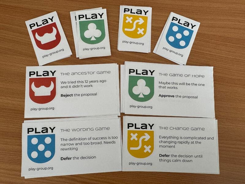 Cards from The Committee Game (from PLaY group): four suit-style badge cards — red "The Ancestor Game" (Reject the proposal — "We tried this 12 years ago and it didn't work"), green "The Game of Hope" (Approve the proposal — "Maybe this will be the one that works"), blue "The Wording Game" (Defer the decision — "The definition of success is too narrow and too broad"), and yellow "The Change Game" (Defer the decision until things calm down — "Everything is complicated and changing rapidly").