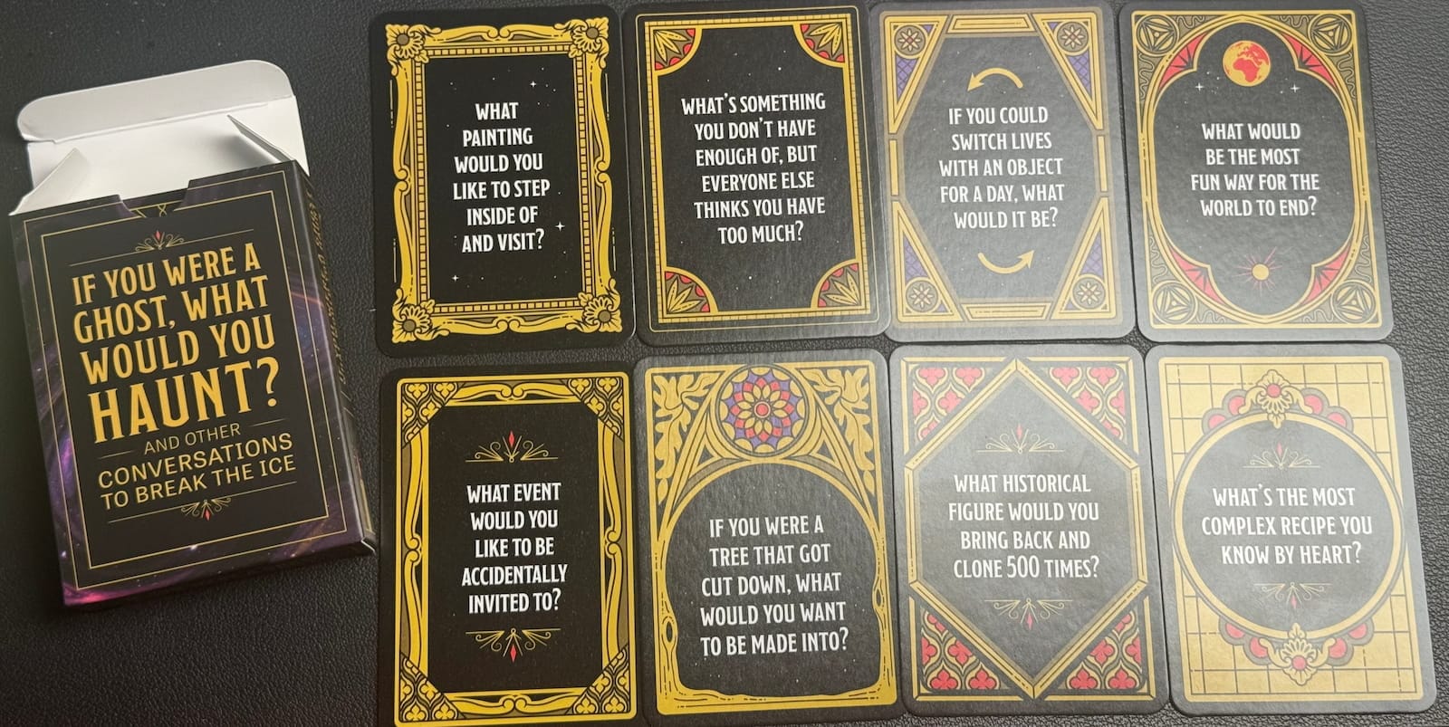 The "If You Were a Ghost, What Would You Haunt? And Other Conversations to Break the Ice" card box beside eight question cards in ornate gold gothic frames, with prompts including "What painting would you like to step inside of and visit?," "If you could switch lives with an object for a day, what would it be?," "What would be the most fun way for the world to end?," and "What historical figure would you bring back and clone 500 times?"
