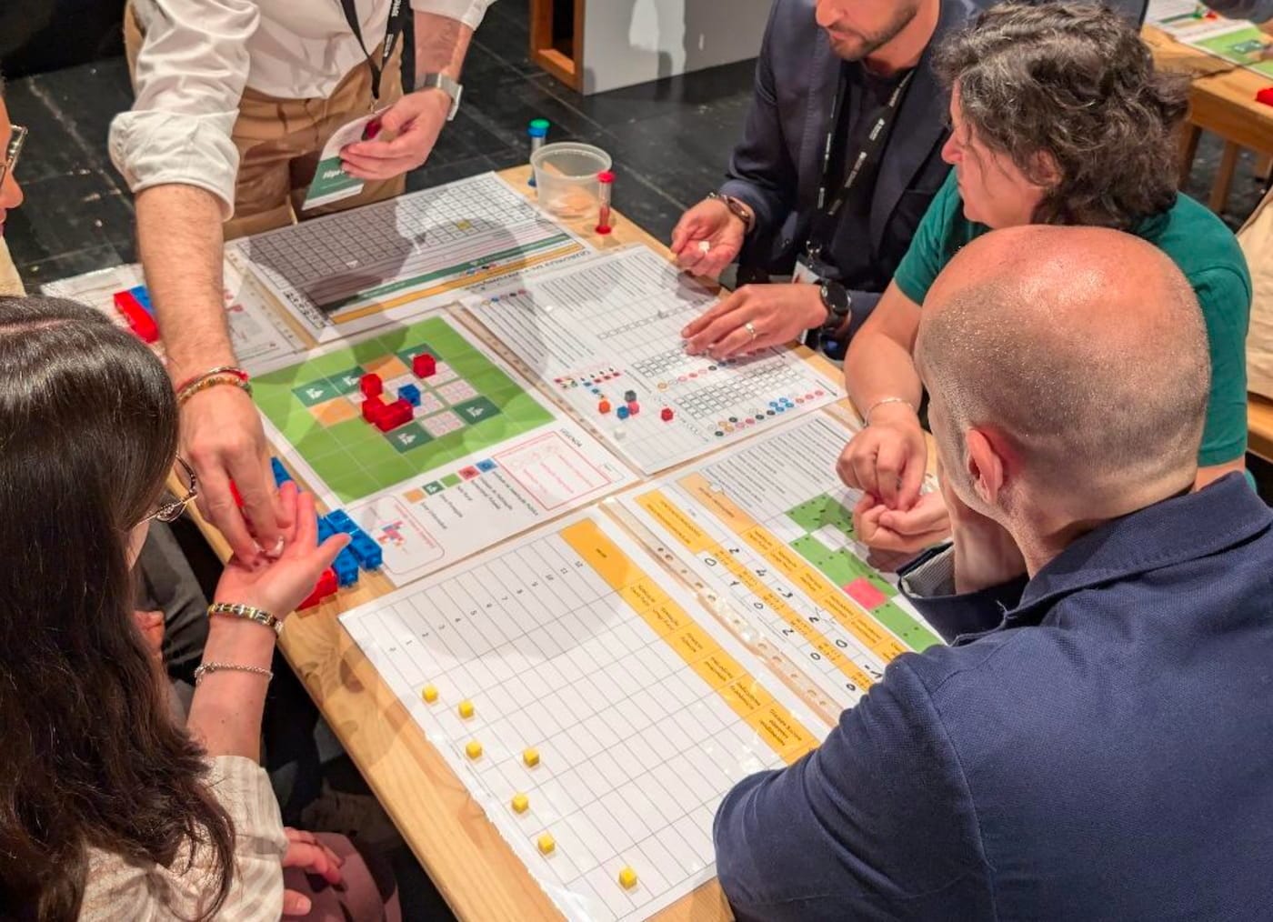 Workshop participants gathered around a table playing Micael Sousa's housing policy game, arranging red and blue building blocks on a gridded map board alongside printed scoring sheets, colored tokens, and geographic visualizations.