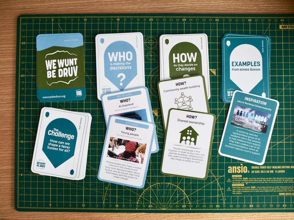 The We Wunt Be Druv card deck laid out on a green cutting mat, showing the category cards — the title card, "The Challenge: How can we shape a fairer Sussex for all?," "Who is making the decisions?," "How do they decide on changes," and "Examples from across Sussex" — alongside sample Who cards ("AI Overlord," "Young people"), How cards ("Community wealth building," "Shared ownership"), and an Inspiration card featuring the Adur & Worthing Climate Assembly.