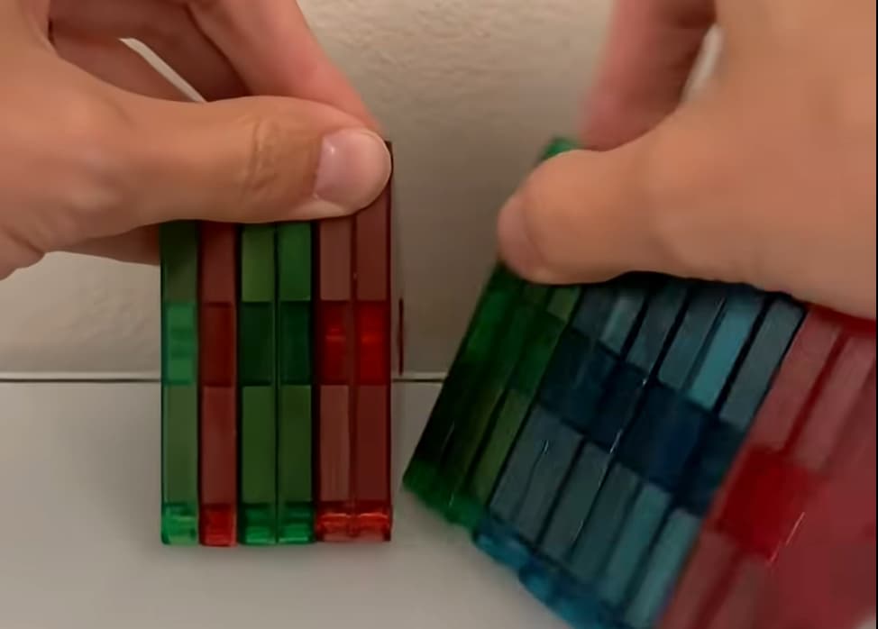 Two hands each holding a tightly packed stack of translucent colored Magna-Tiles — greens and reds grouped on the left, blues, greens, and reds on the right — demonstrating the sorting-and-grouping move behind the clustering algorithm.