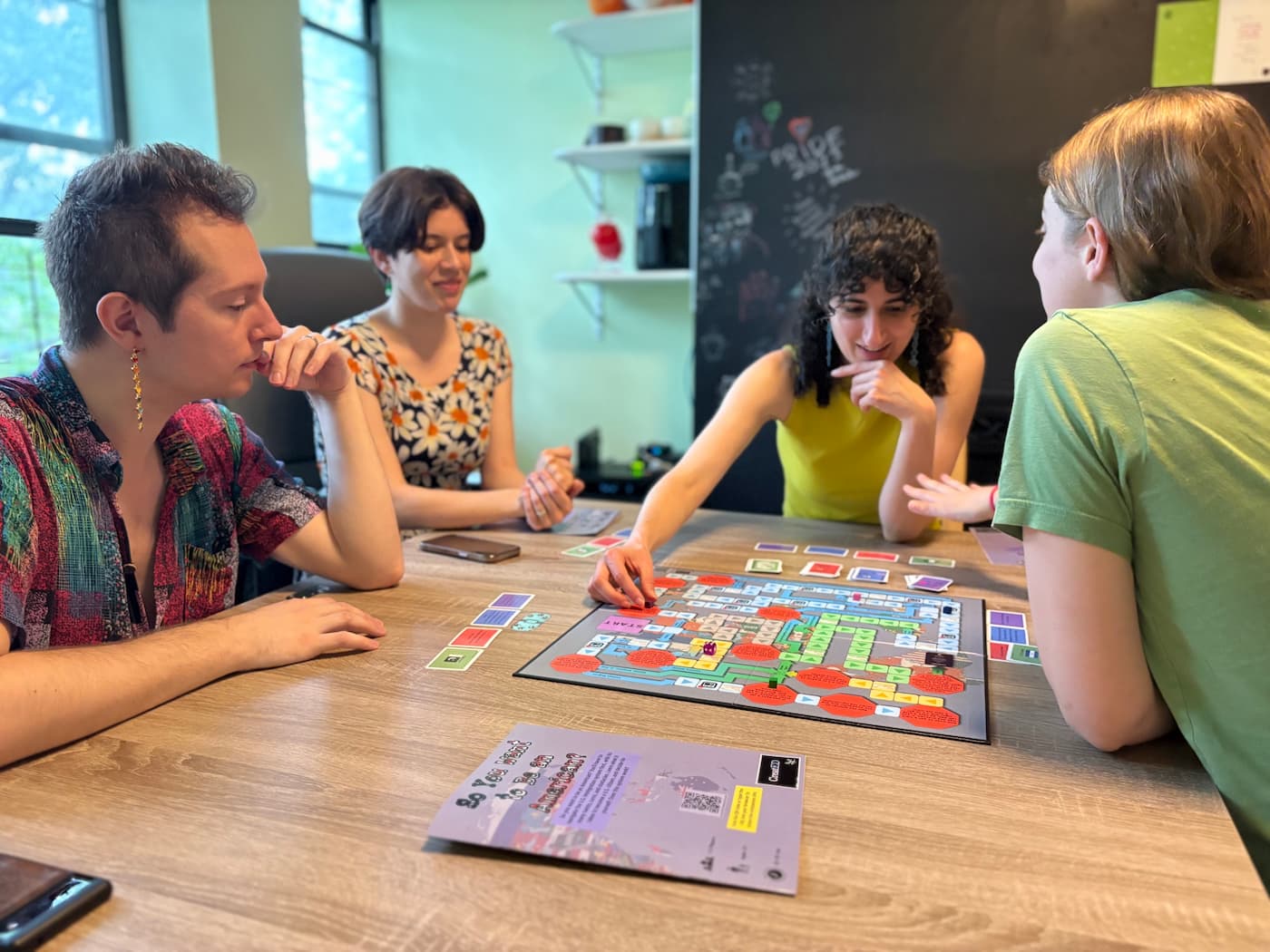 Four people seated around a wooden table playing the analog board game "So You Want to Be an American?", with a colorful tiled board, small cards, and dice mid-play.