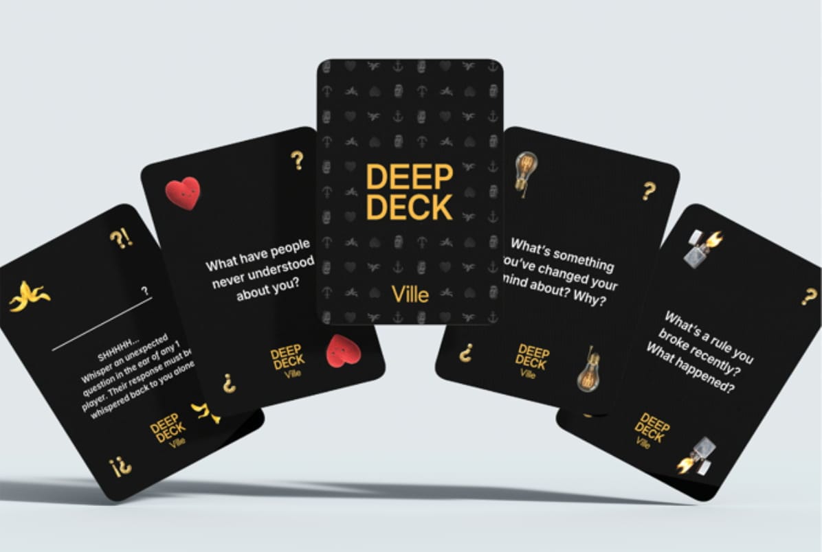 The Deep Deck (by Ville) fanned out against a pale background: black cards with gold and white type. The center card shows the "DEEP DECK" logo over a patterned back of small suit-like symbols (anchor, microphone, lightbulb, heart, banana, and more). Surrounding question cards read "What have people never understood about you?", "What's something you've changed your mind about? Why?", and "What's a rule you broke recently? What happened?", each marked with a symbol in the corner; a left-hand action card gives a whispered-question instruction.