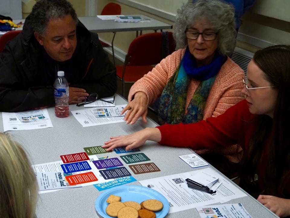 Four people seated around a table at a community screening, leaning in and pointing at a spread of colored discussion cards and printed handouts, with a plate of biscuits to the side.