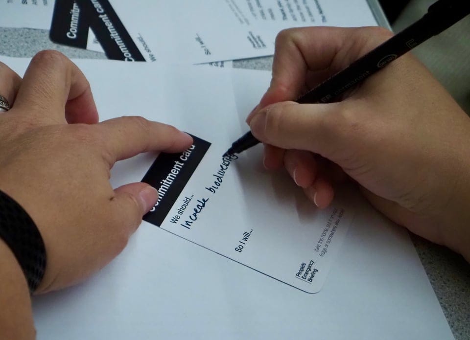 Close-up of two people at a table; one writes with a marker on a “Commitment card” printed with the prompts “We should…” and “So I will…,” adding a handwritten response. More People’s Emergency Briefing cards lie nearby.