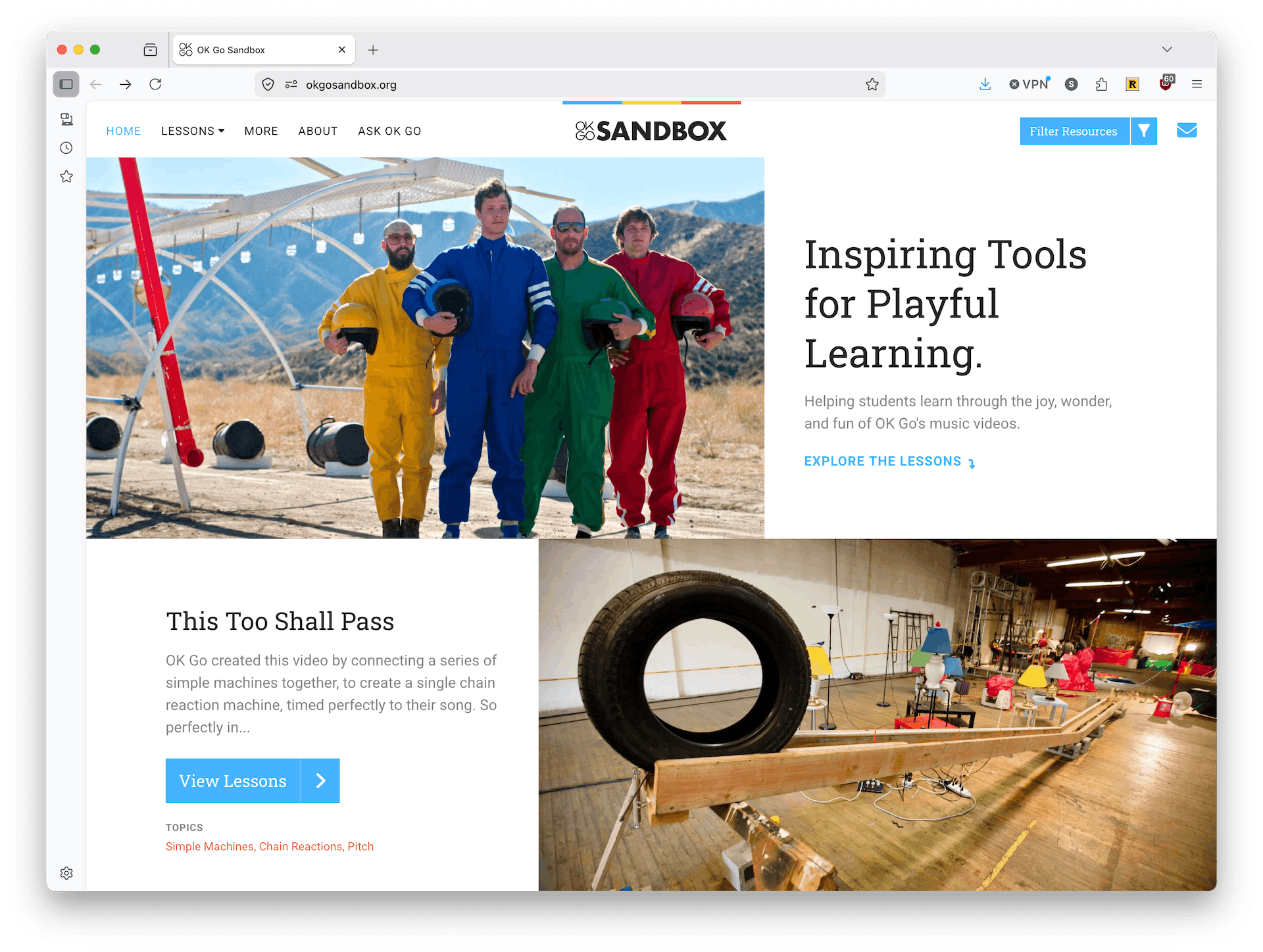 Browser window on okgosandbox.org. The hero shows the four members of OK Go in colored jumpsuits (yellow, blue, green, red) holding helmets in front of a desert rig, beside the heading "Inspiring Tools for Playful Learning" and the line "Helping students learn through the joy, wonder, and fun of OK Go's music videos." Below, a "This Too Shall Pass" lesson card sits next to a photo of the band's Rube Goldberg-style chain-reaction machine (a tire rolling down a wooden ramp in a warehouse), tagged with topics Simple Machines, Chain Reactions, and Pitch.