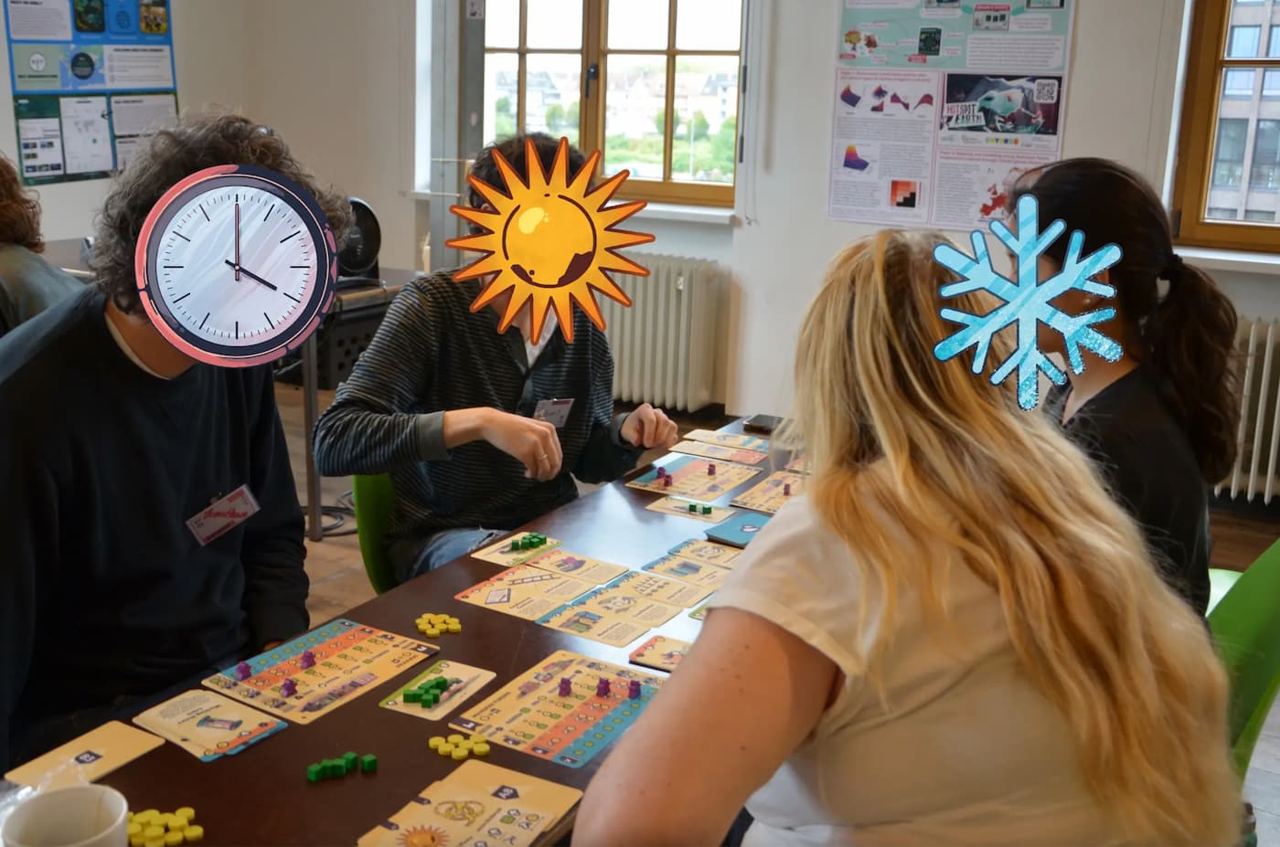Four people playing the cooperative board game Izdihar around a table covered with illustrated player boards, cards, and colored wooden cubes; their faces are hidden behind cartoon clock, sun, and snowflake stickers. Research posters hang on the wall behind them.