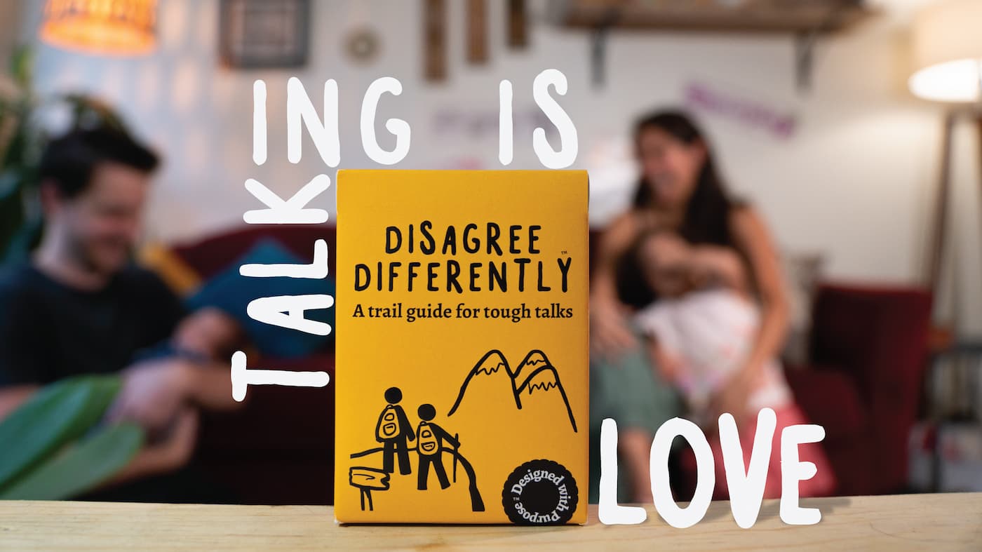 The Disagree Differently box standing on a table — yellow, with a hand-drawn illustration of two hikers climbing toward a summit, the tagline “A trail guide for tough talks,” and a “Designed with Purpose” badge. Large white letters spelling “TALKING IS LOVE” frame the scene, with a family softly blurred in the background.